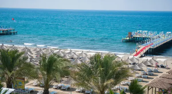 Sonne, Strand und Meer am quattro beach spa & resort. Liegestühle unter Strohschirmen, Palmen und ein langer Steg mit rotem Teppich laden zum Entspannen ein.