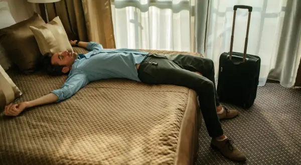 Erschöpfter Mann liegt auf einem Hotelbett, Koffer daneben. Der Jetlag von Asien nach Deutschland hat ihn völlig ausgeknockt.