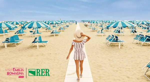 Frau mit Hut geht auf einem Weg am Strand von Rimini. Hunderte von Liegestühlen und Sonnenschirmen warten auf den perfekten Rimini Urlaub.