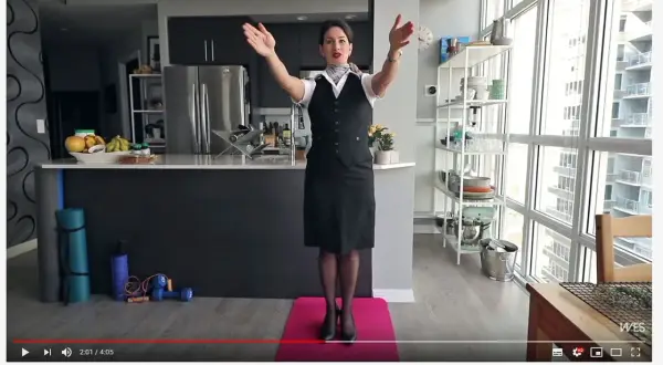 Eine Stewardess oder Flugbegleiterin in Uniform steht auf einer rosa Matte und macht Übungen. Im Hintergrund eine Küche und ein großes Fenster.