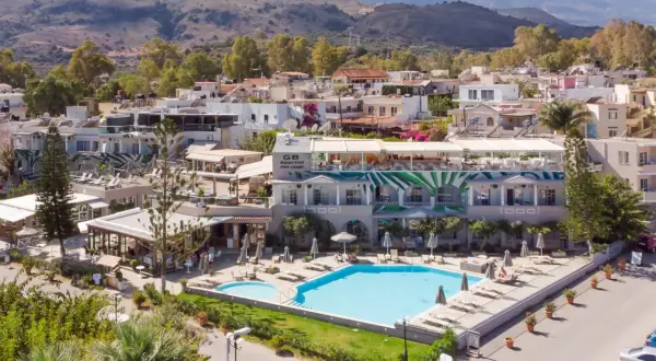 Ein **epos luxury beach hotel** mit Pool und Liegestühlen, umgeben von mediterraner Vegetation und Häusern, im Hintergrund Berge.