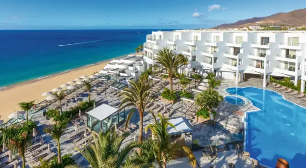 Traumhafter Blick auf das Riu Palace Jandia: Weißes Hotel, Pool, Palmen und der goldene Strand am türkisfarbenen Meer.