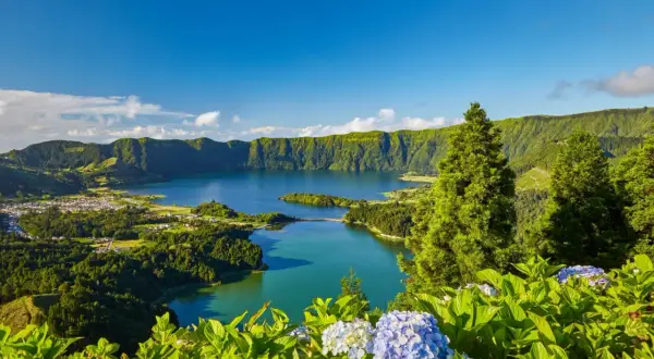 Grüne Hügel umrahmen einen tiefblauen See. Hier liegen die Azoren, eine Inselgruppe im Atlantik, mit üppiger Vegetation und blühenden Hortensien im Vordergrund.