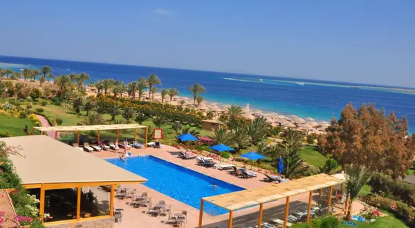Ein luxuriöser Poolbereich mit Blick auf das Meer und den Strand des Fort Arabesque West Bay. Palmen und Sonnenschirme säumen den Weg zum türkisfarbenen Wasser.