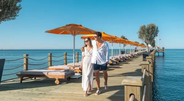 Ein Paar spaziert über einen Holzsteg am Meer, gesäumt von Liegen und Sonnenschirmen. Ein perfekter Urlaub im Arum Barut Collection.