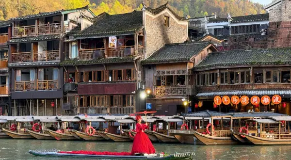 Ein Urlaub in China: Frau in rotem Kleid auf Boot vor traditionellen Häusern und Booten am Fluss.