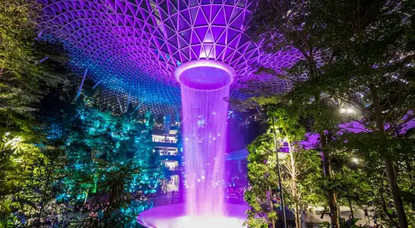 Imposanter Wasserfall im Jewel Changi Airport Singapur, beleuchtet in lila und blau unter einem geometrischen Glasdach.