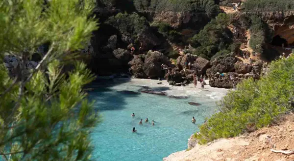 Türkisblaues Wasser in einer malerischen Bucht auf Mallorca. Menschen sonnen sich auf Felsen und schwimmen im Meer.