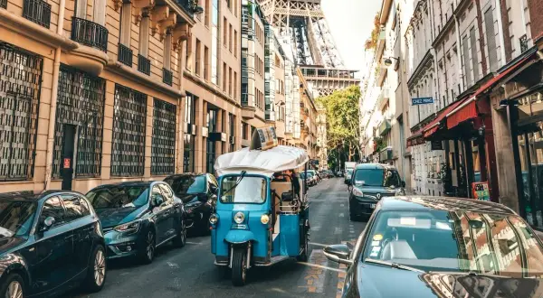Ein Tuk-Tuk fährt durch Paris, vorbei an Autos und Gebäuden, mit dem Eiffelturm im Hintergrund. Die Kosten für eine Fahrt mit dem Auto nach Paris sind hier nicht ersichtlich.
