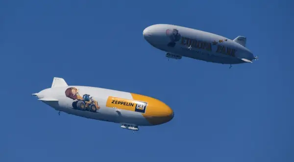 Zwei Zeppeline, einer mit einem Bagger und "Zeppelin CAT", der andere mit "Europa Park" und einem Koala, schweben am blauen Himmel.