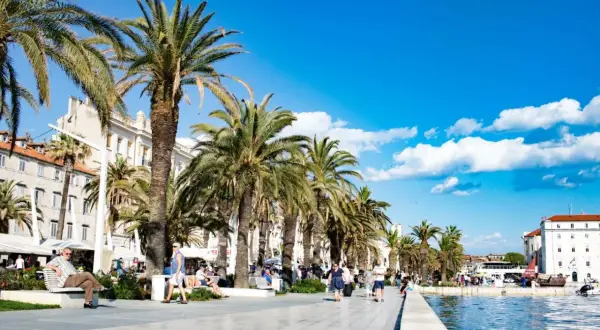Palmen säumen die Uferpromenade in Split, Kroatien. Menschen spazieren und entspannen sich unter blauem Himmel.