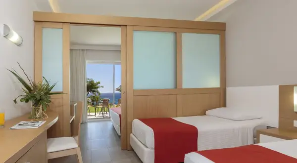 Zwei Betten mit roten Decken im Rodos Princess Beach Hotel. Ein Schreibtisch mit Blumen und Blick aufs Meer.