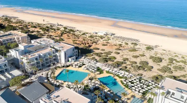 Luftaufnahme des Hipotels Barrosa Park mit Pools, Liegestühlen und Palmen, daneben ein breiter Sandstrand und das blaue Meer.