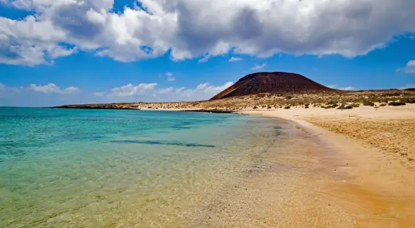 Traumhafter Strand mit türkisfarbenem Wasser und einem Vulkan im Hintergrund – ideal für Ihren Kanaren Urlaub.