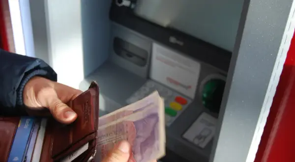 Person hält Geld und Portemonnaie vor Geldautomat. Hier kann man bei der Bank Geld wechseln.