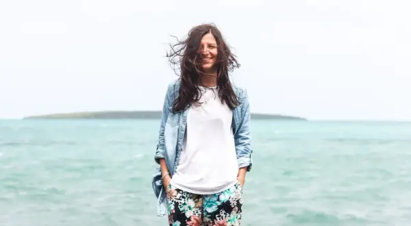 Frau mit wehendem Haar am Meer, die lächelt. Perfekt für die Packliste Mauritius: leichte Kleidung, Jeansjacke und bunte Hose.