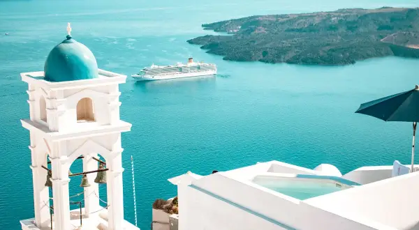 Traumhafter Urlaub Griechenland: Weißer Glockenturm mit blauer Kuppel, Kreuzfahrtschiff auf dem Meer, Infinity-Pool und Sonnenschirm.