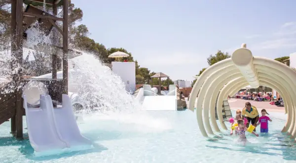 Kinder planschen im Wasserpark, während ein Wasserfall vom Rutschenturm herabstürzt. Ein Mann mit gelbem Shirt beaufsichtigt die Kleinen unter einem knochenartigen Tunnel in den Calimera Fido Gardens.