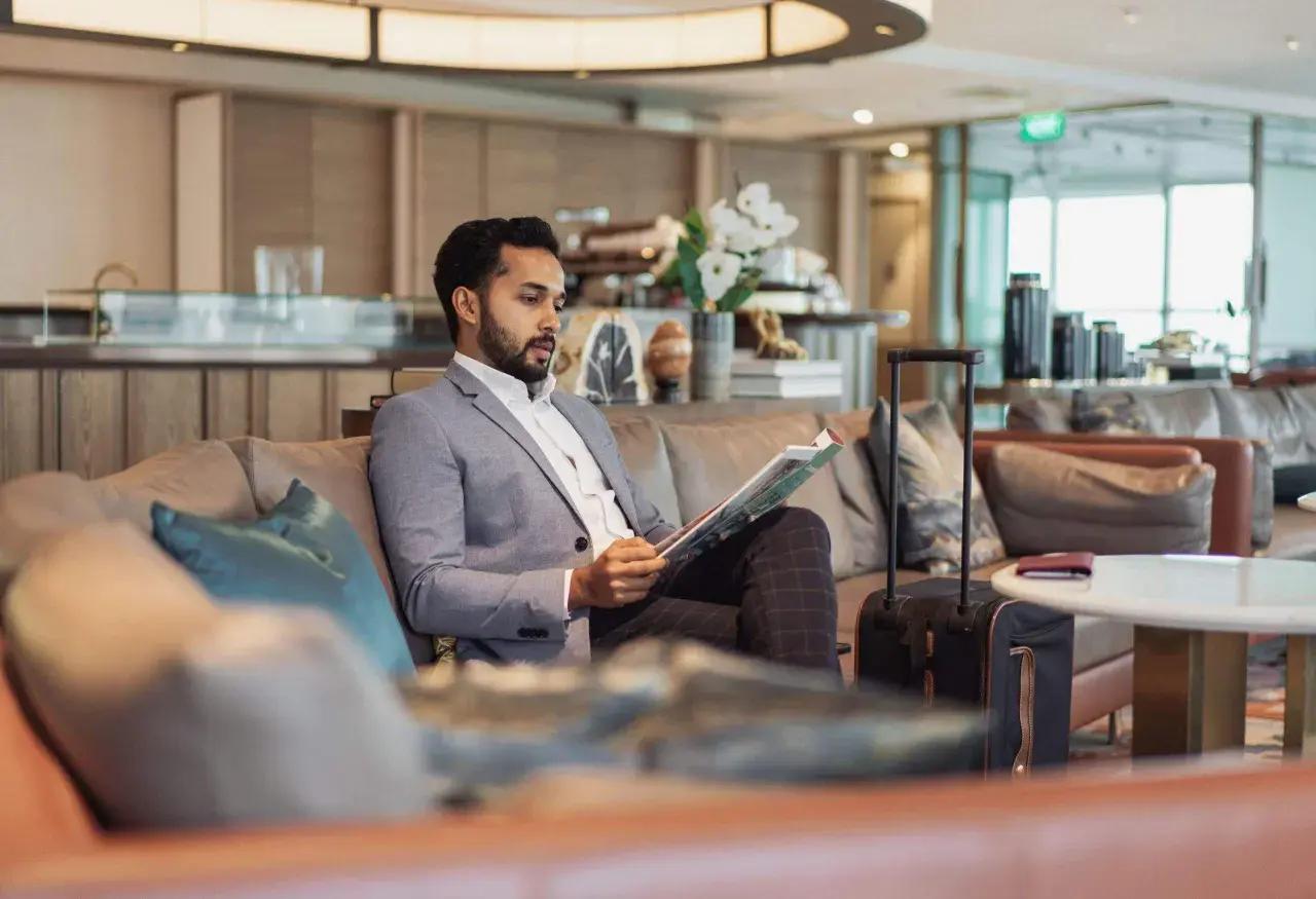 Mann im Anzug liest Zeitschrift in der airport VIP lounge. Sein Koffer steht bereit.