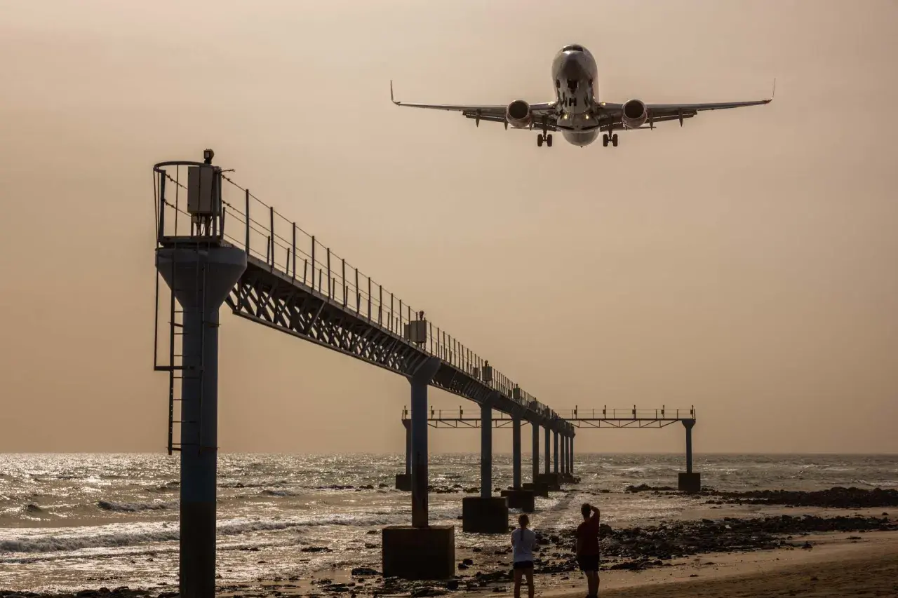 Zwei Personen genießen das Plane Spotting am Strand, während ein Flugzeug über eine Seebrücke hinwegfliegt.