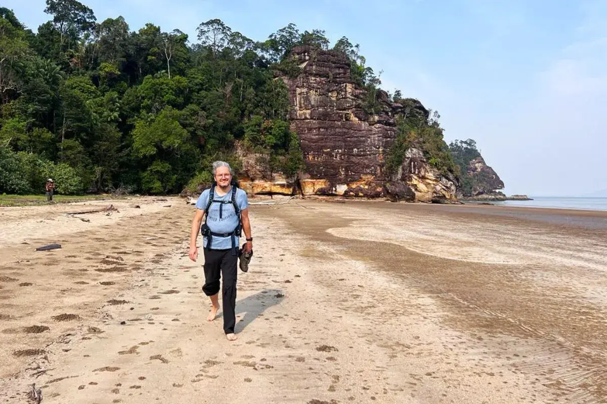 Mann mit Rucksack und Schuhen in der Hand geht barfuß am Strand entlang. Die Packliste für Malaysia ist komplett, die Natur ist üppig.