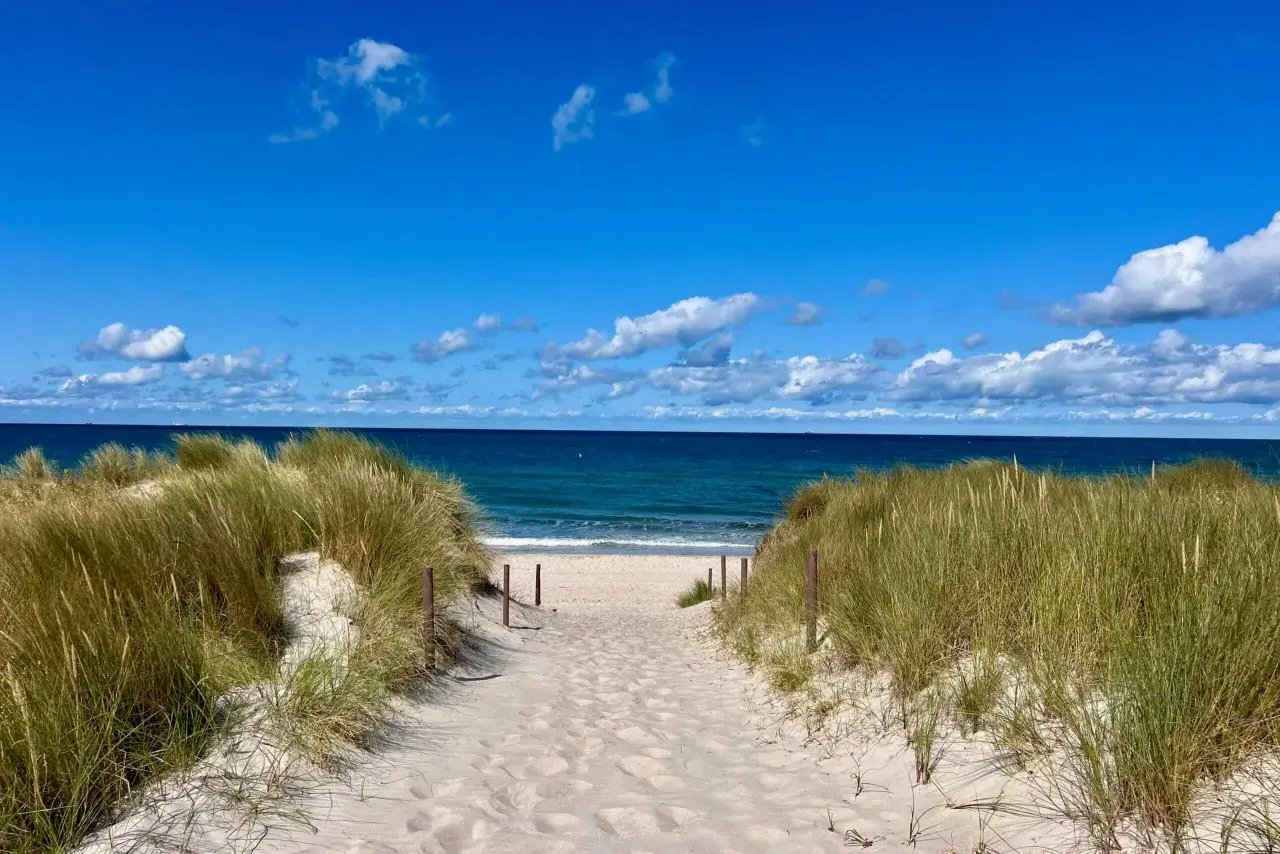 Sandiger Weg durch Dünen zum blauen Meer. Perfekter Urlaub am Meer mit Sonnenschein und Wolken.