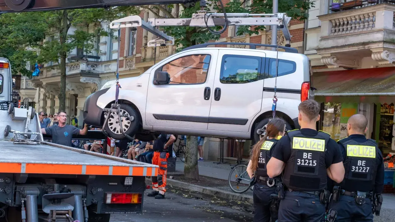 Polizei schleppt ein weißes Auto ab. Ein Mann in orangefarbener Arbeitskleidung hilft. Die auto abschleppen kosten sind hier wohl nicht das Thema.