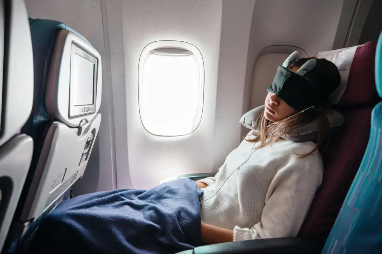 Frau schläft im Flugzeug, mit Schlafmaske und Nackenkissen, eingewickelt in eine Decke.