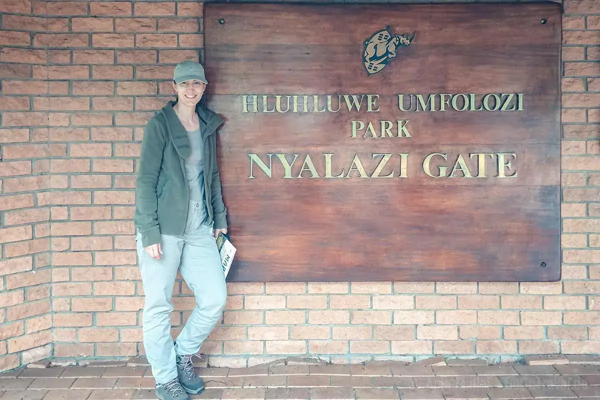 Frau am Eingangstor des Hluhluwe Umfolozi Parks, bereit für ihr Safari-Abenteuer. Ihre Kleidung ist perfekt für die Packliste.