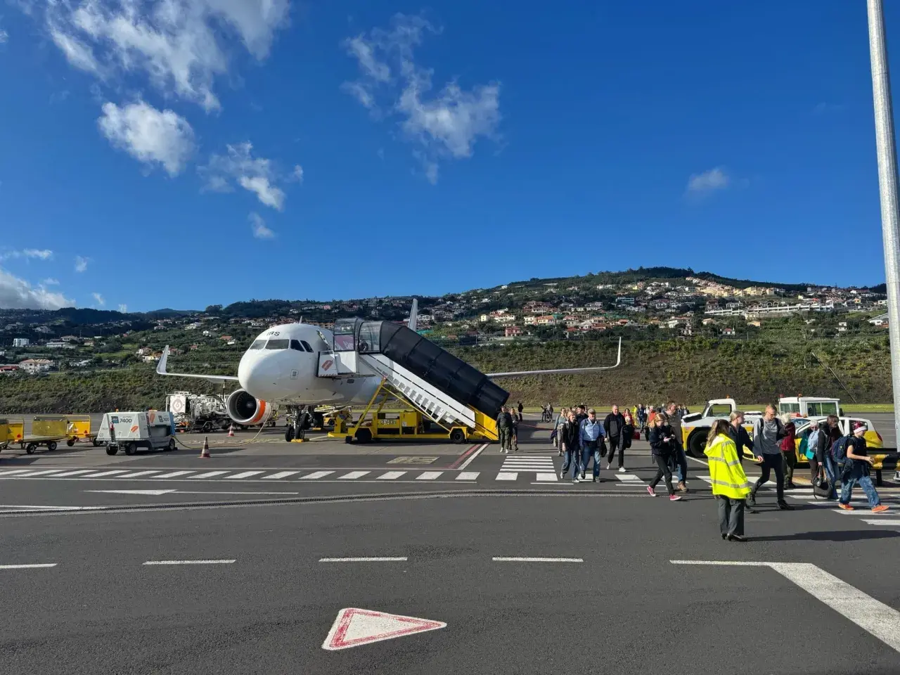 Passagiere steigen am Madeira Flughafen aus dem Flugzeug. Im Hintergrund erstreckt sich eine hügelige Landschaft mit Häusern.