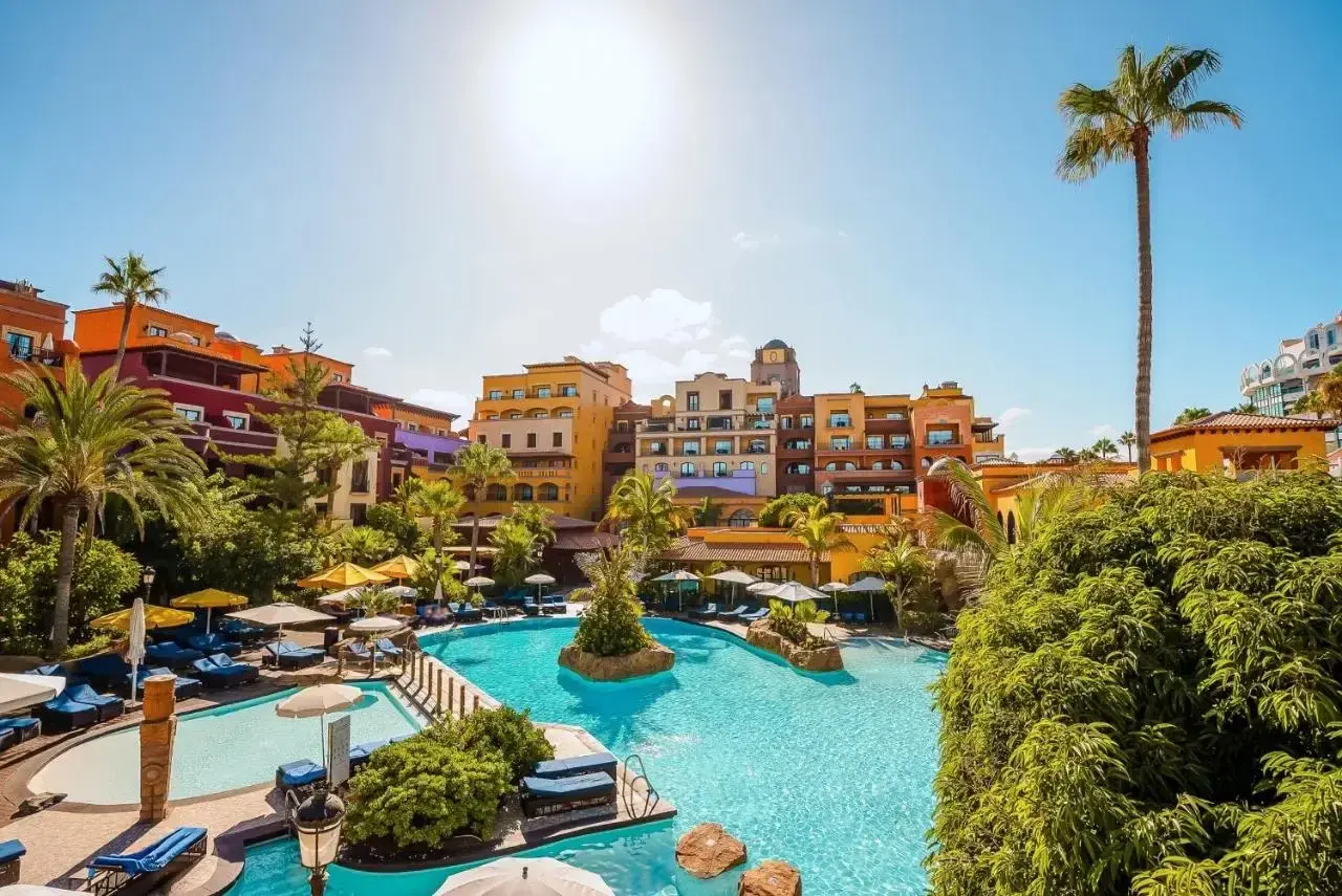 Sonnige Poolanlage mit bunten Gebäuden und Palmen im Riu Palace Teneriffa. Entspannung pur unter blauem Himmel.