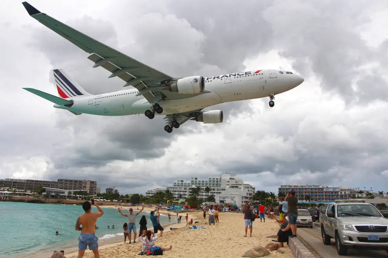 Air France Flugzeug landet auf einem Flughafen direkt am Strand. Menschen amüsieren sich am Meer, während das Flugzeug über sie hinwegfliegt.