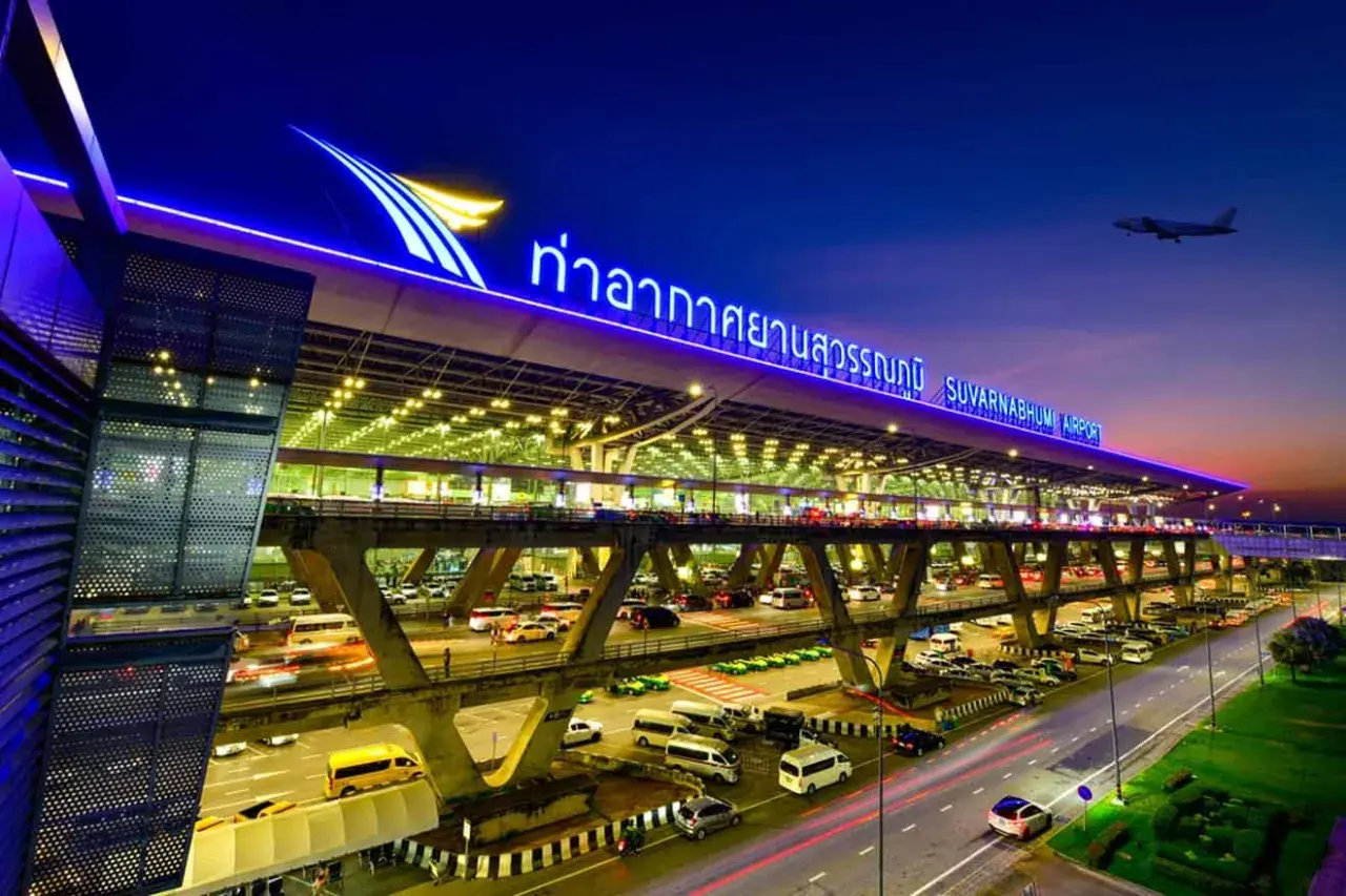 Der beleuchtete Flughafen Bangkok Suvarnabhumi bei Nacht, mit Autos auf mehrstöckigen Zufahrtsstraßen und einem Flugzeug am Himmel.