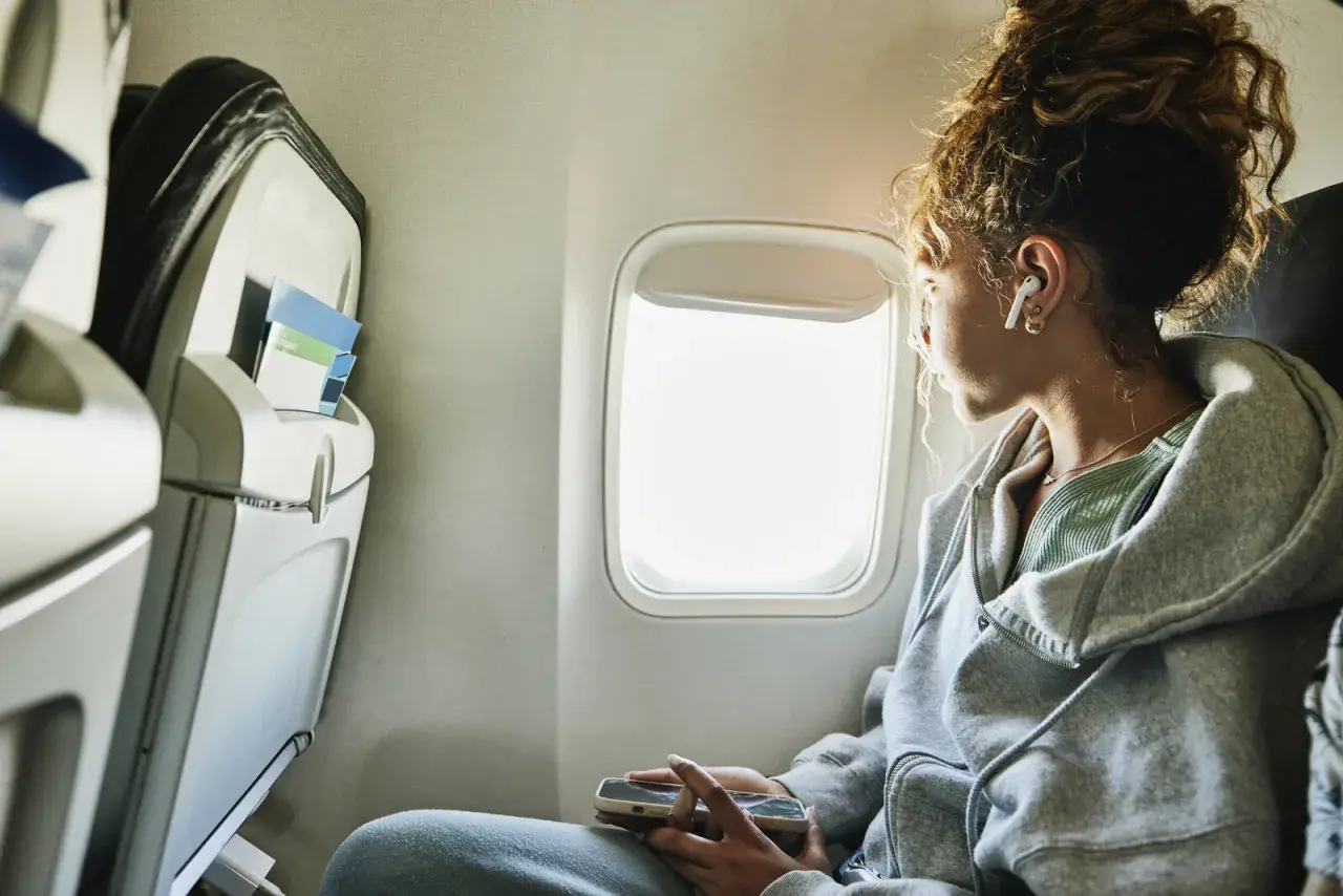 Junge Frau blickt aus dem Flugzeugfenster, vertieft in ihr Handy. Sie fragt sich, wie ist es zu fliegen, während sie die Wolken vorbeiziehen sieht.