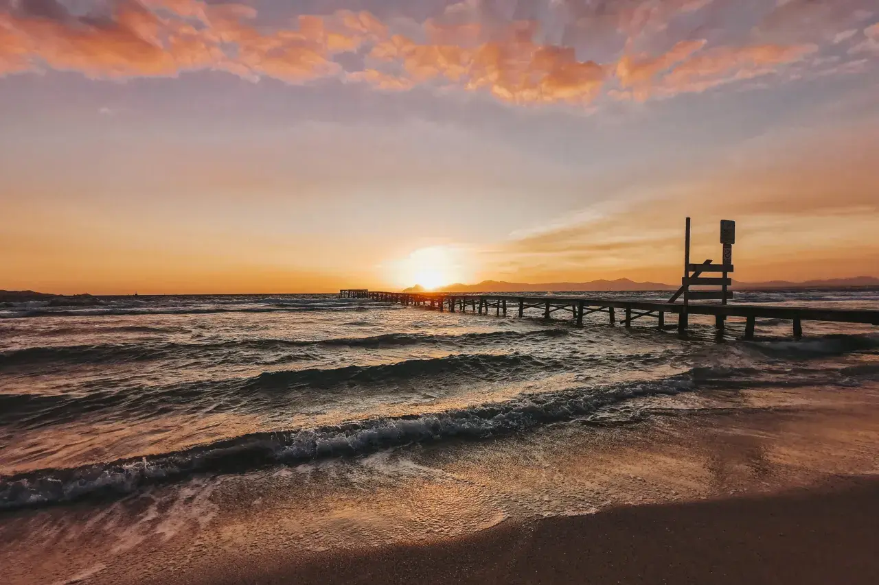 Sonnenuntergang über dem Meer an der Playa de Muro. Wellen brechen sanft am Strand, ein langer Holzsteg führt ins Wasser.