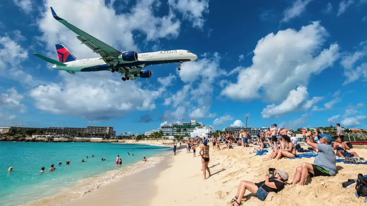 Ein Delta-Flugzeug landet tief über einem belebten Strand in der Karibik. Urlauber am **Karibik Flughafen** genießen die Sonne, während das Flugzeug über ihnen kreist.
