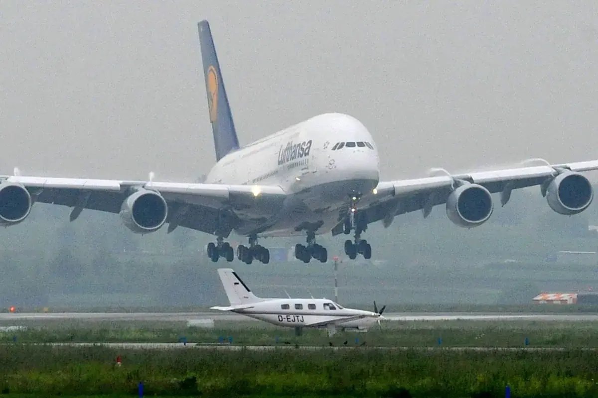Ein riesiger A380 von Lufthansa landet, sein enormes Gewicht wird durch die vier Triebwerke und das Fahrwerk sichtbar. Ein kleines Propellerflugzeug wartet daneben.