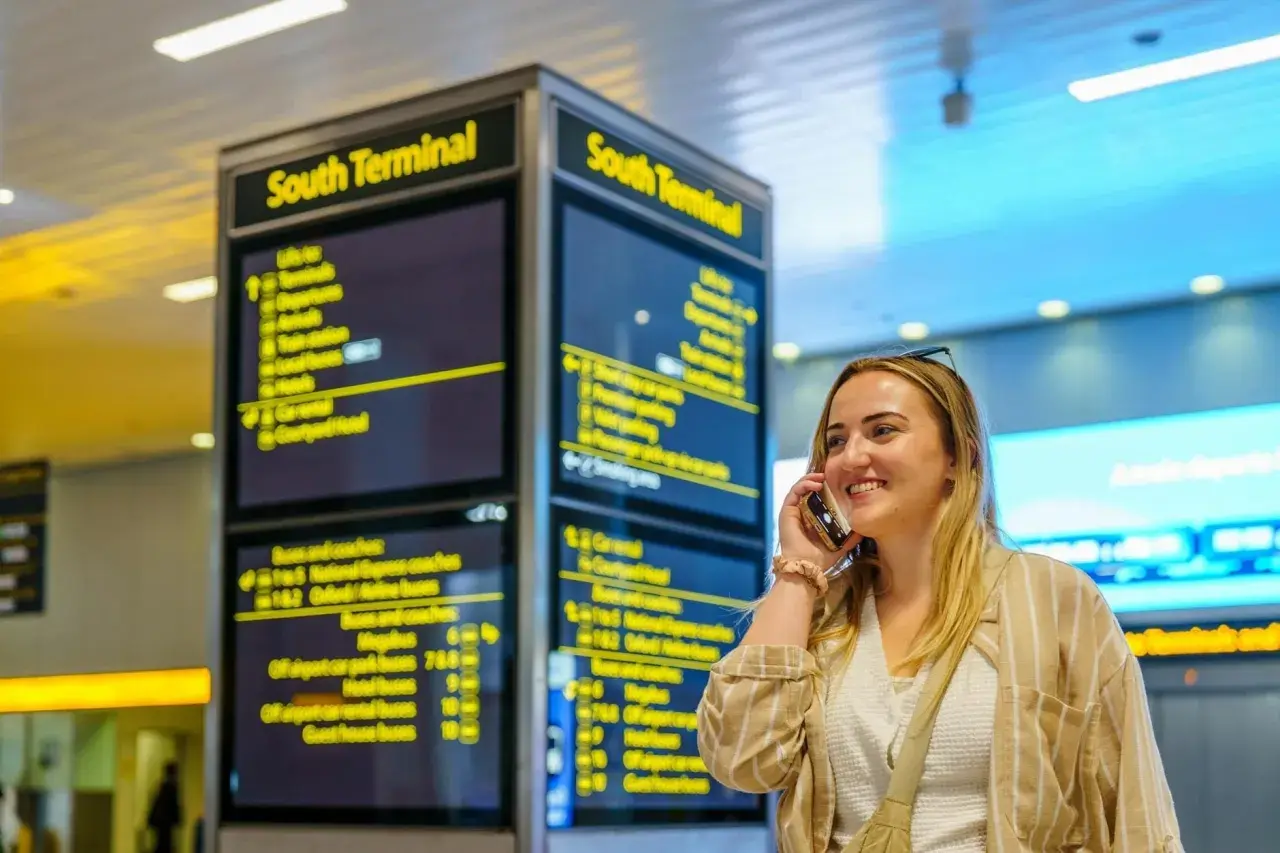 Frau telefoniert am London Flughafen vor einer Anzeigetafel für den South Terminal.