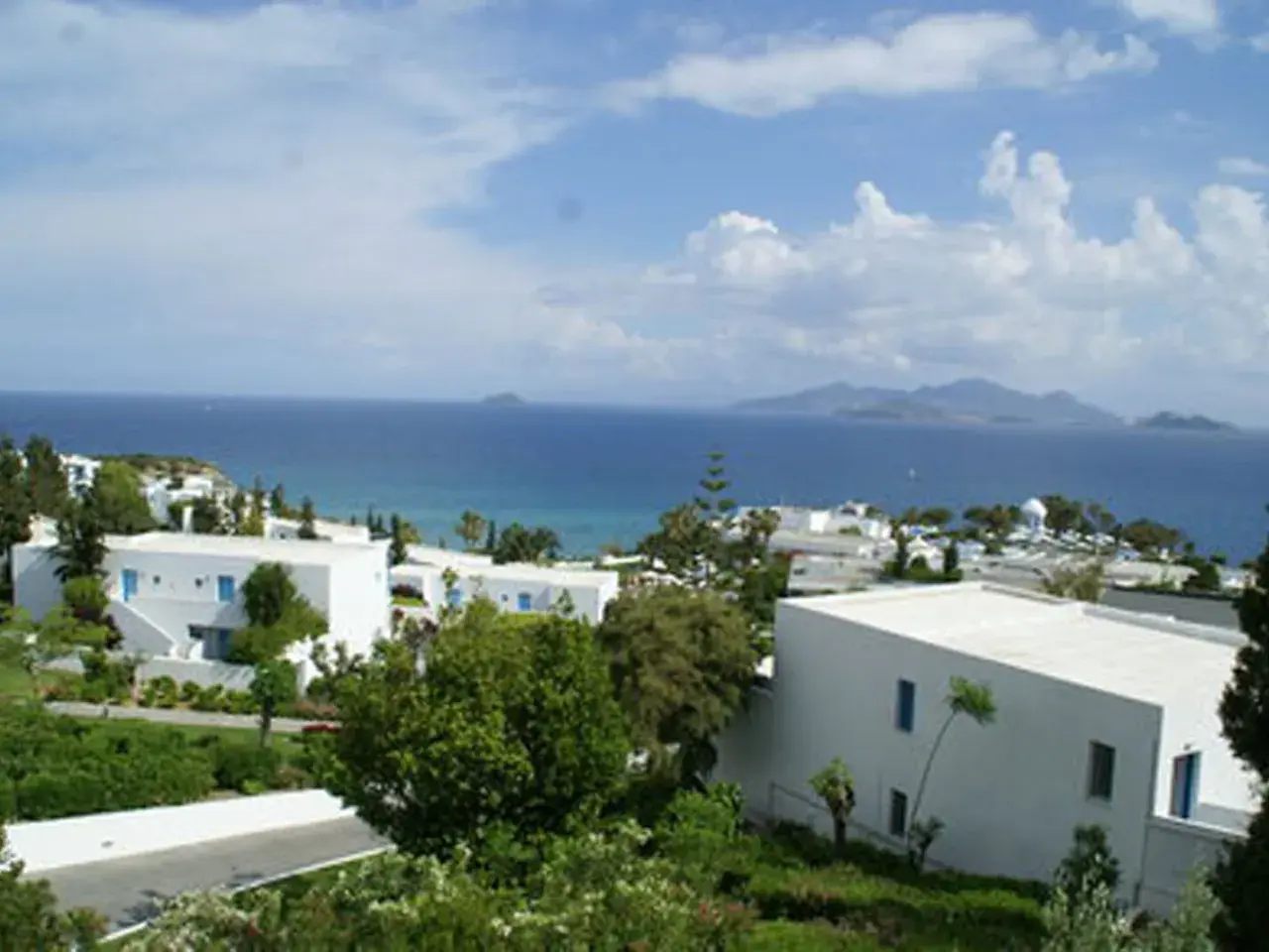 Blick auf das Robinson Daidalos Resort mit weißen Gebäuden und blauen Fenstern, umgeben von grüner Vegetation, am Meer mit Inseln am Horizont.