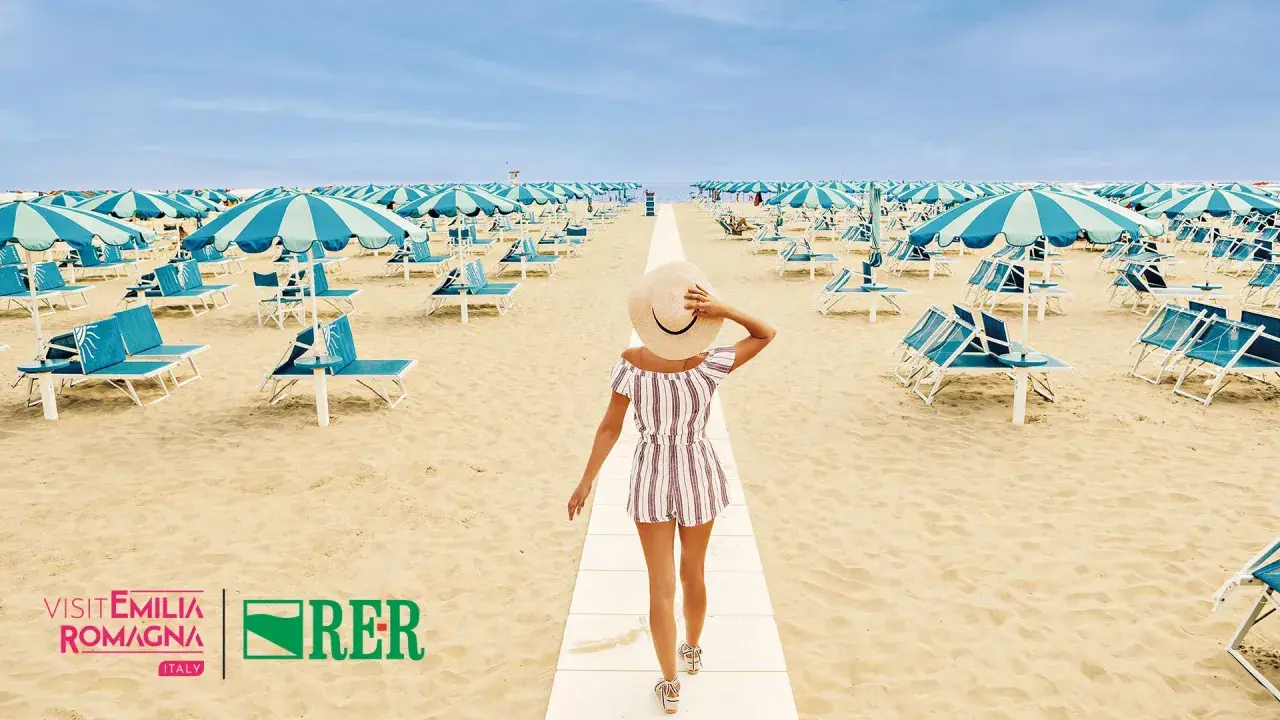 Frau mit Hut geht auf einem Weg am Strand von Rimini. Hunderte von Liegestühlen und Sonnenschirmen warten auf den perfekten Rimini Urlaub.