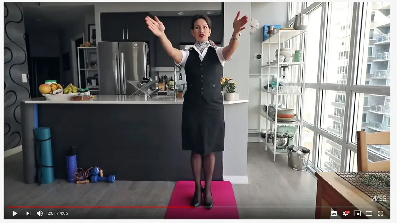Eine Stewardess oder Flugbegleiterin in Uniform steht auf einer rosa Matte und macht Übungen. Im Hintergrund eine Küche und ein großes Fenster.