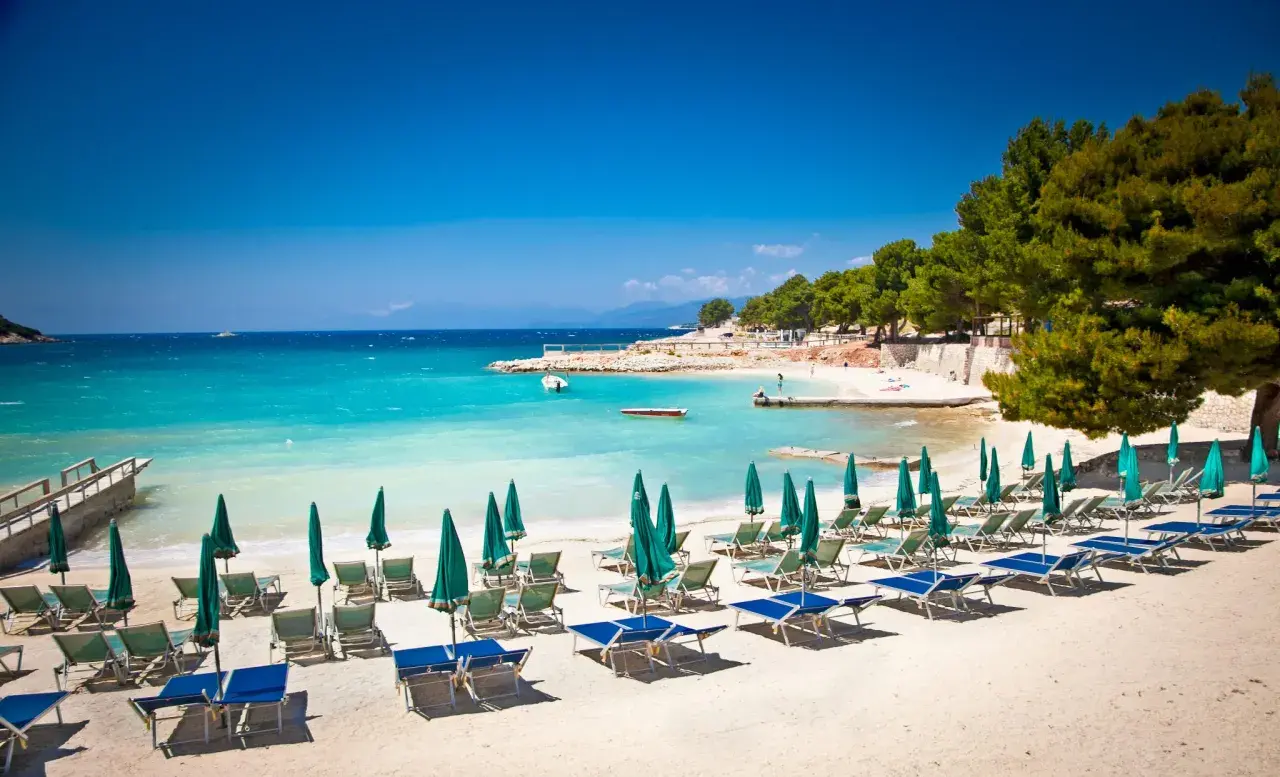Ein sonniger **Tirana Strand** mit türkisfarbenem Wasser, Liegestühlen und Sonnenschirmen.