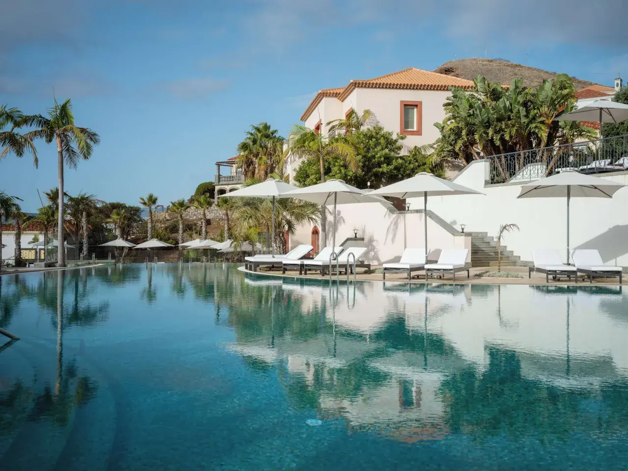 Traumhafter Poolbereich mit Liegen und Sonnenschirmen im Dreams Madeira Spa & Marina. Palmen und ein elegantes Gebäude im Hintergrund.
