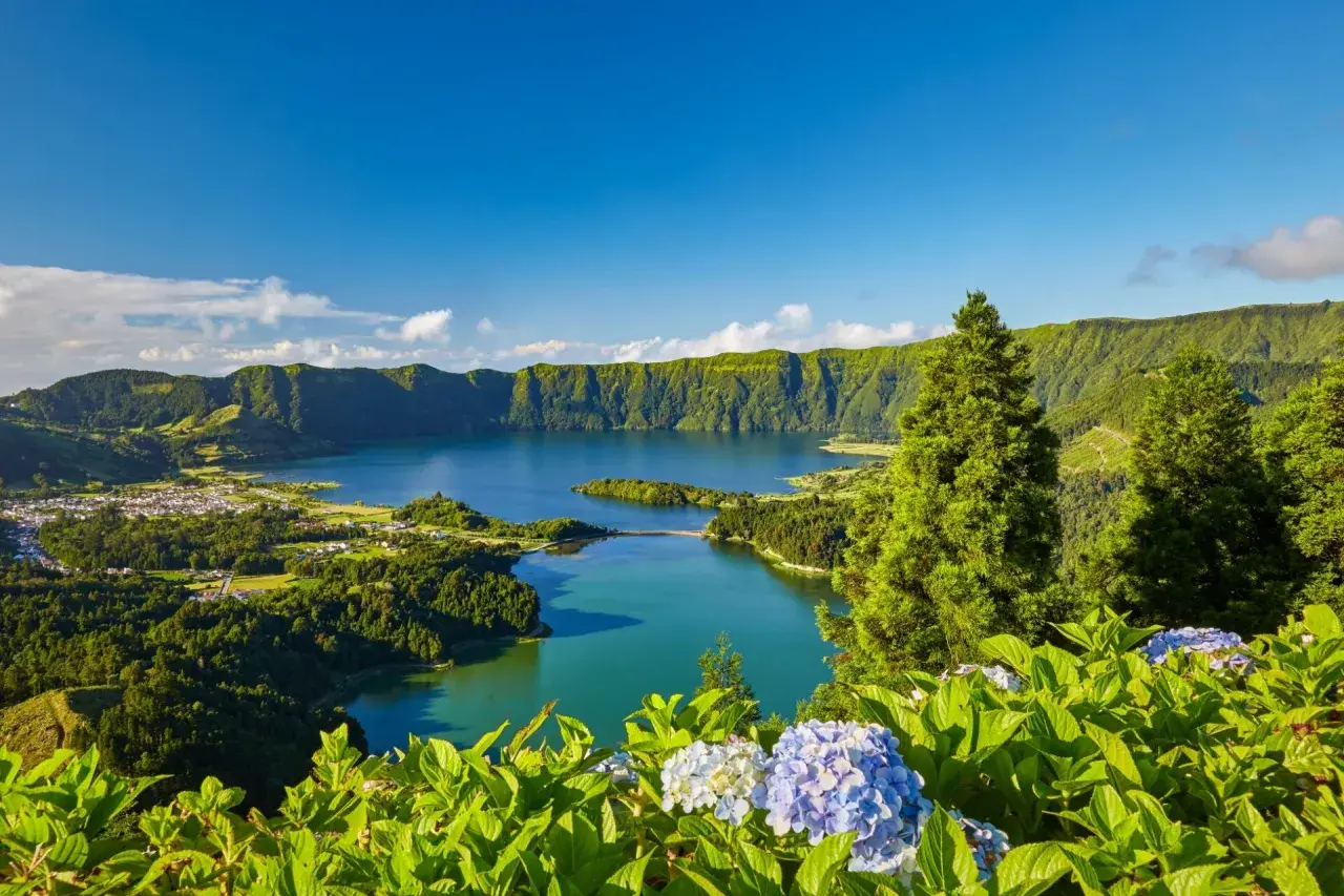 Grüne Hügel umrahmen einen tiefblauen See. Hier liegen die Azoren, eine Inselgruppe im Atlantik, mit üppiger Vegetation und blühenden Hortensien im Vordergrund.