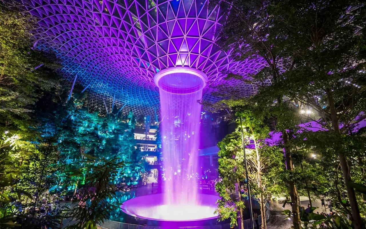 Imposanter Wasserfall im Jewel Changi Airport Singapur, beleuchtet in lila und blau unter einem geometrischen Glasdach.
