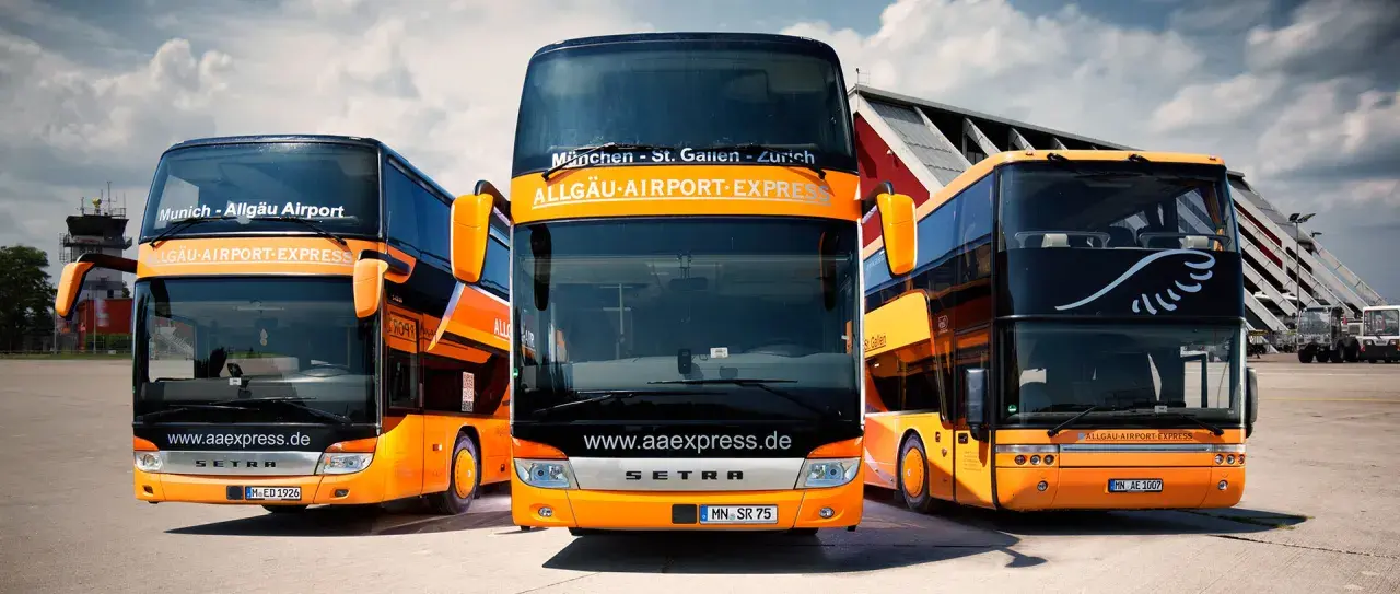 Drei orange-schwarze Doppeldeckerbusse von Allgäu Airport Express stehen bereit. Informieren Sie sich über die alex airport shuttle preise für Ihre Reise.