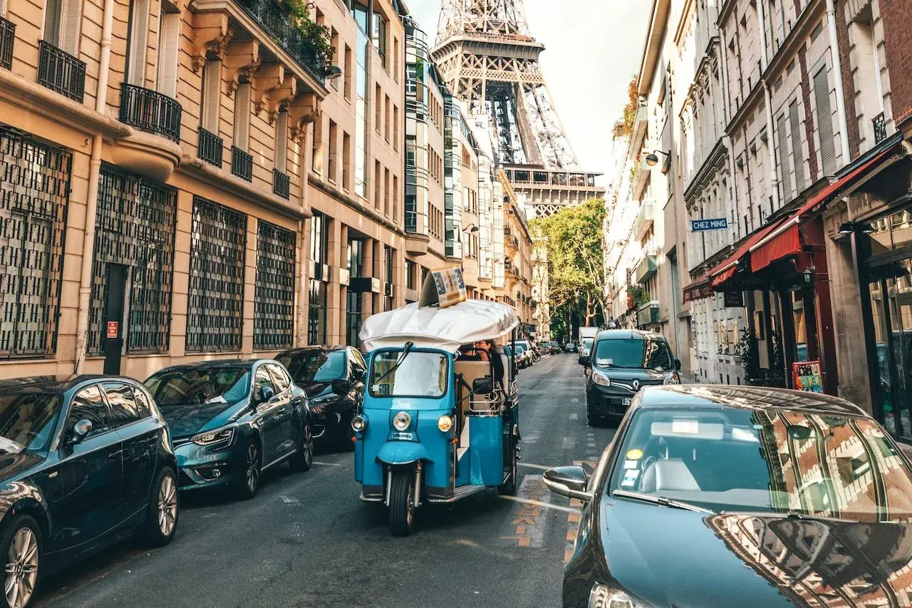 Ein Tuk-Tuk fährt durch Paris, vorbei an Autos und Gebäuden, mit dem Eiffelturm im Hintergrund. Die Kosten für eine Fahrt mit dem Auto nach Paris sind hier nicht ersichtlich.