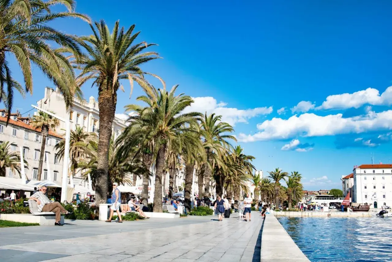Palmen säumen die Uferpromenade in Split, Kroatien. Menschen spazieren und entspannen sich unter blauem Himmel.