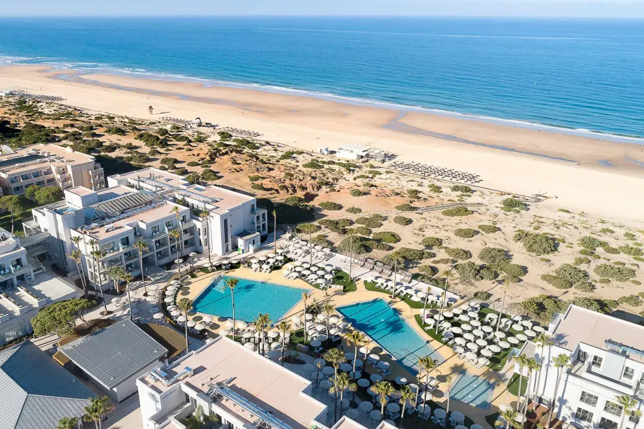 Luftaufnahme des Hipotels Barrosa Park mit Pools, Liegestühlen und Palmen, daneben ein breiter Sandstrand und das blaue Meer.