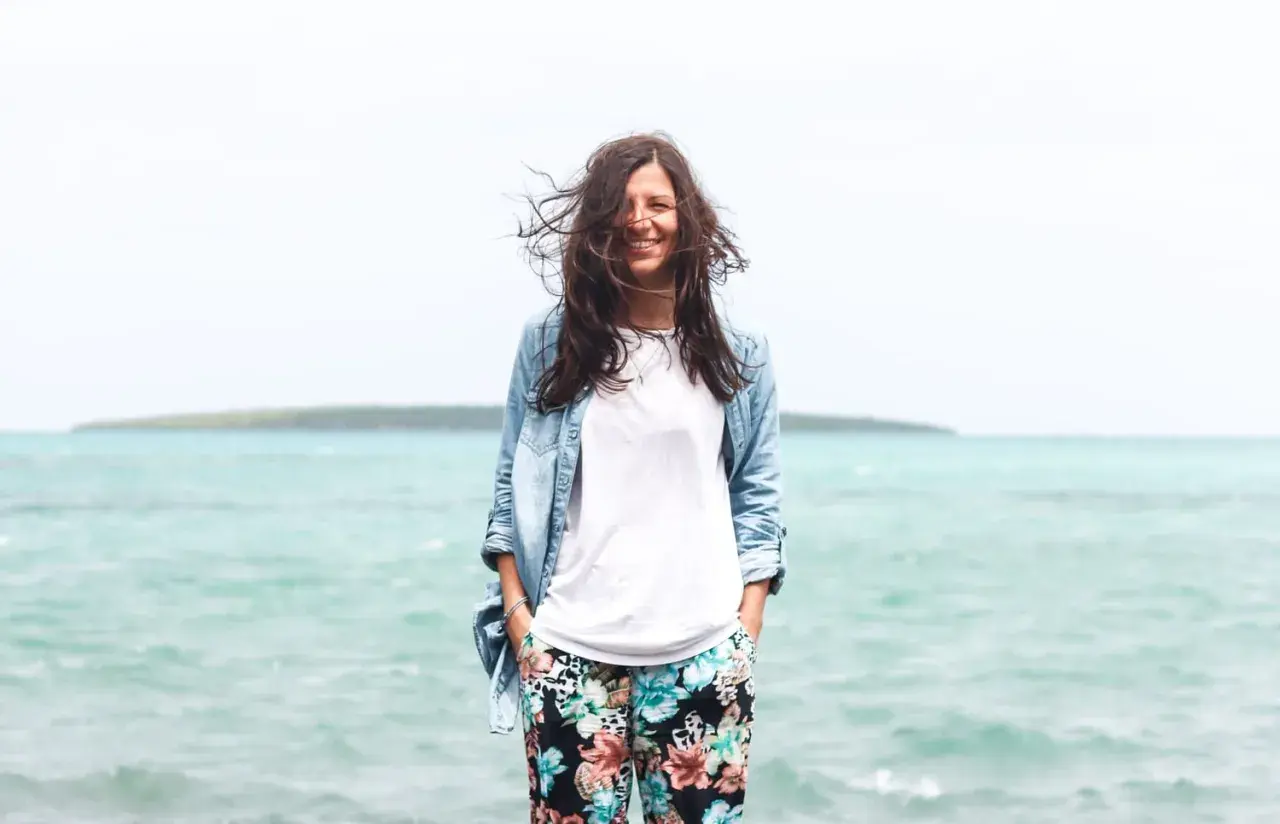 Frau mit wehendem Haar am Meer, die lächelt. Perfekt für die Packliste Mauritius: leichte Kleidung, Jeansjacke und bunte Hose.
